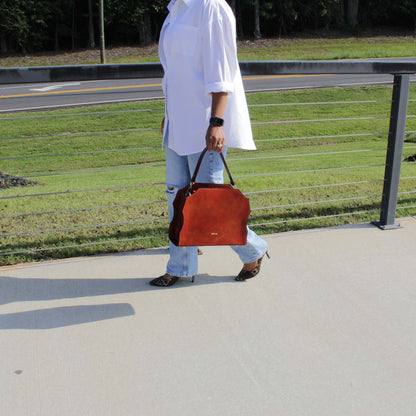 Hand‑painted full‑grain leather Forgiveness structured satchel handcrafted in cognac, held by a woman in motion by the top handle in a lifestyle view.  